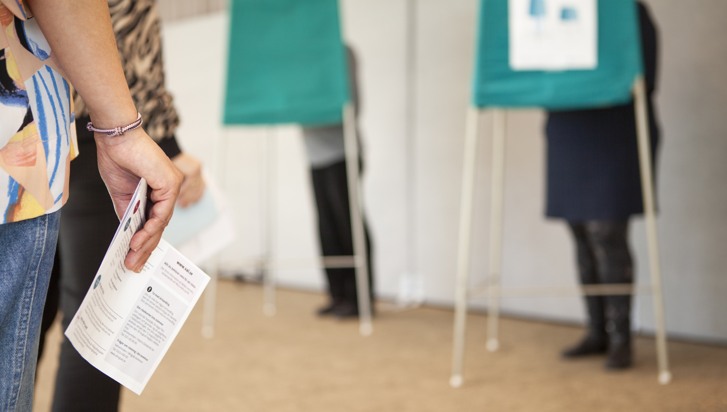 Människor i kö med röstkort i händerna som väntar på att rösta bakom avskilda skärmar.