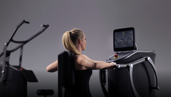 En kvinna sitter vid en träningsmaskin som är kopplad till e-gym. På displayen framför sig ser hon grafik som visar i vilken takt hon ska genomföra övningen.