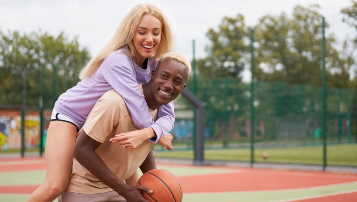 Två unga personer skrattar tillsammans. En kvinna hoppar upp på ryggen av en man som håller i en basketboll
