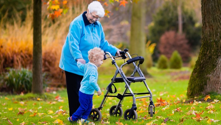 Äldre kvinna med rollator på promenad i parken med litet barn.