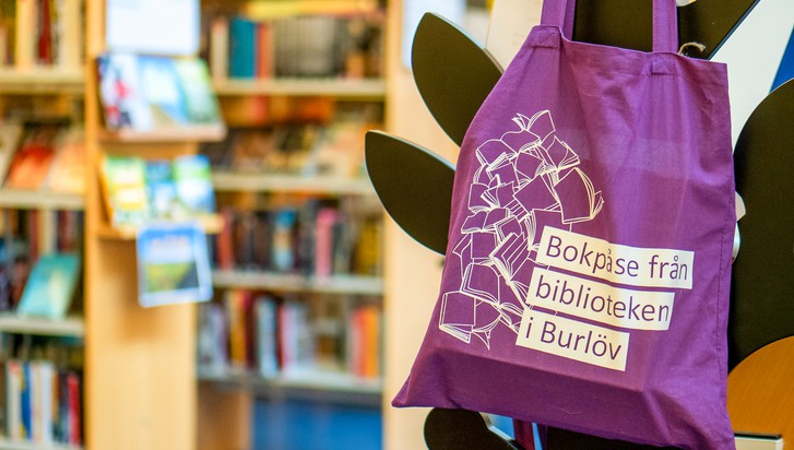 lila tygpåse med texten "Bokpåse från biblioteken i Burlöv" hänger på en klädhängare med bokhyllor i bakgrunden