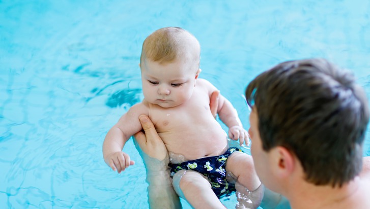 En glad medelålders man badar med sin bebis i en pool.